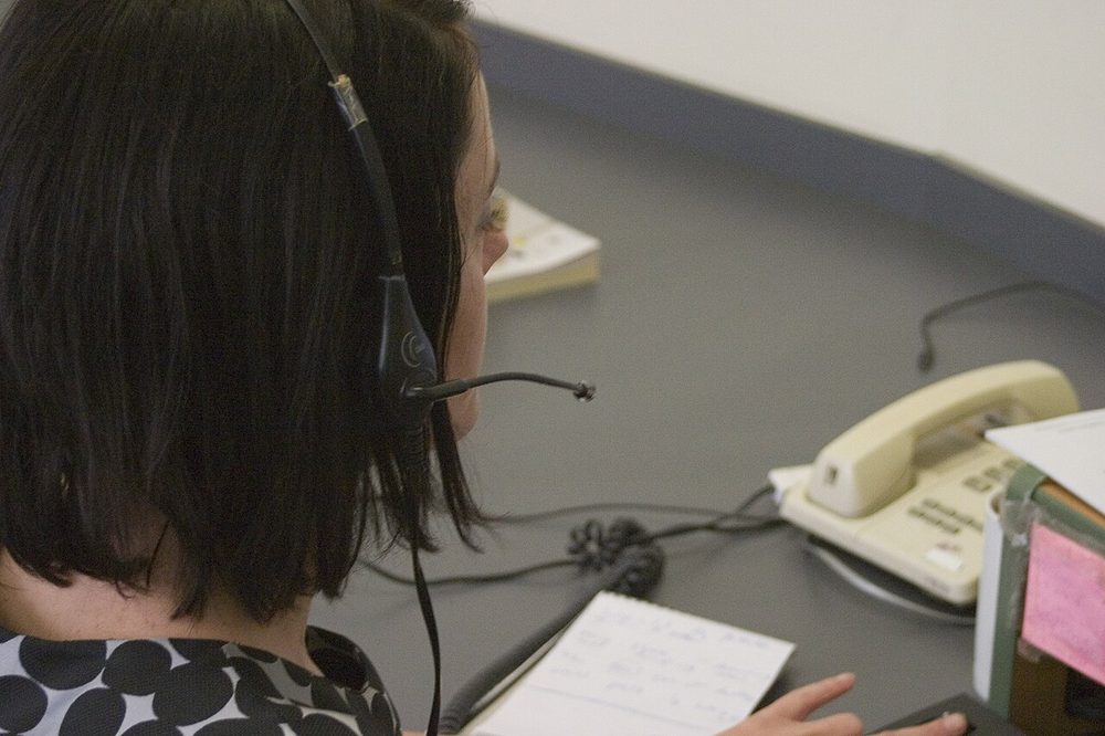 A contact-centre agent wearing a telephone headset at a desk beside a desk phone and notepad