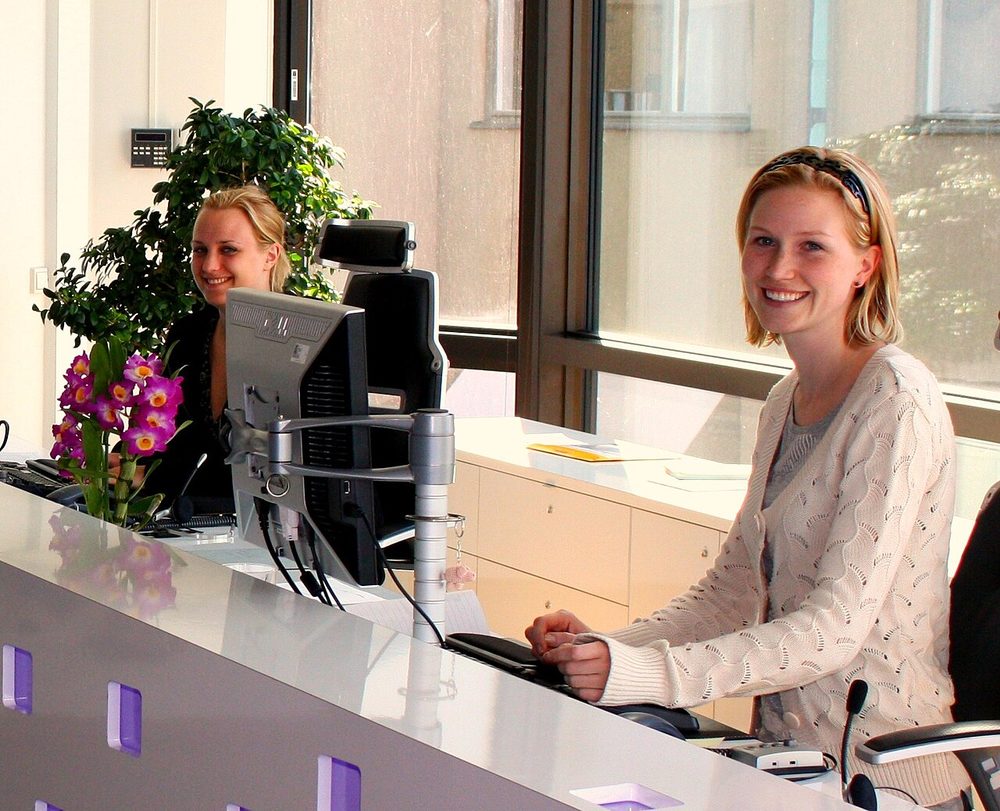 Two smiling receptionists at a bright front desk with computer monitors and a desk microphone
